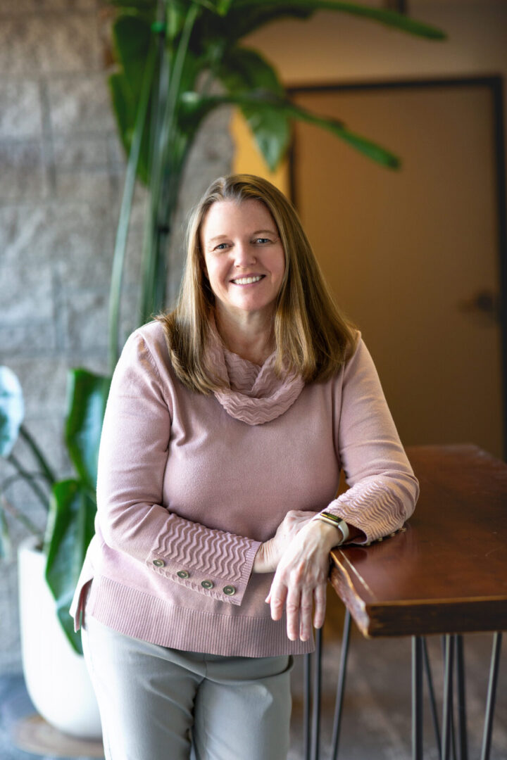 Smiling woman in a cozy pink sweater sitting indoors by a wooden table.